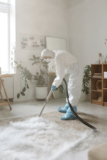 A young boy using a portable vacuum cleaner to deep clean a beige carpet in a living room while an older man, likely a family member, is resting on a white sofa nearby. The room features light gray wa